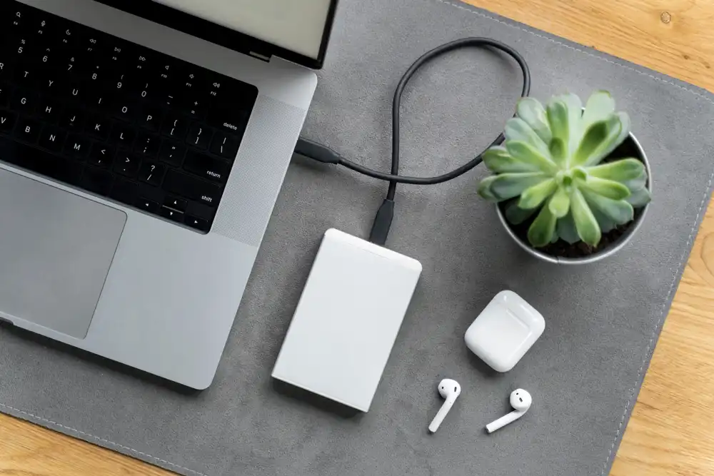 External hard drive capacity - معرفی بهترین هارد اکسترنال 2 ترابایت در سال 1402 هارد اکسترنال سفید روی میز طوسی متصل به لپ تاپ طوسی در کنار ایر پاد سفید و گلدان سفید کاکتوس